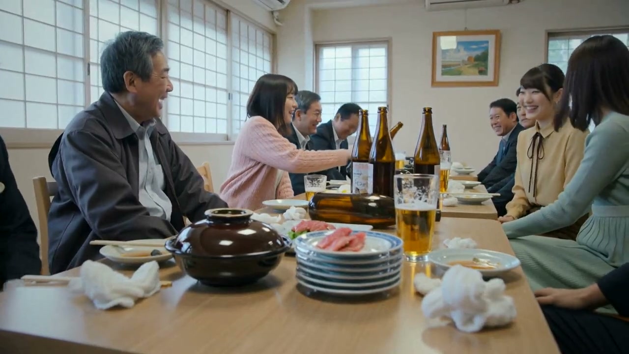 座敷の宴会。卓に土鍋と刺身の大皿、ビール瓶が並び、参加者が談笑している